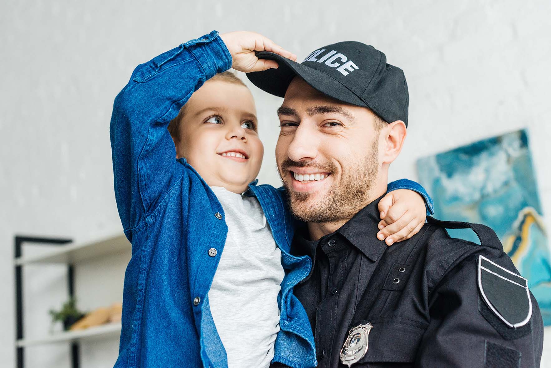 Close-up portrait of smiling young father in police uniform carrying his little son and looking at camera.