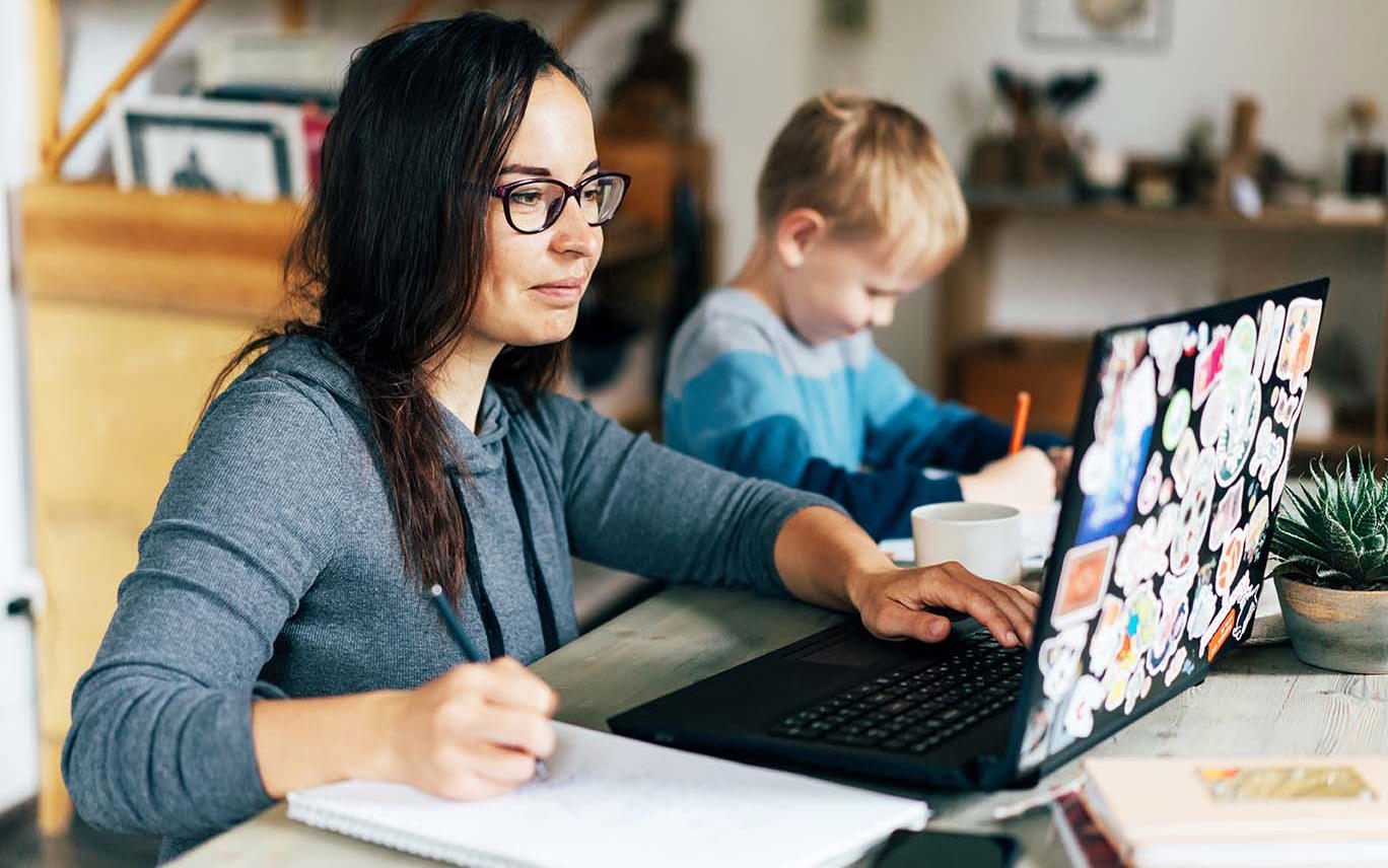 Concept of work from home and home family education. Mom and son are sitting at the desk. Business woman works on the Internet in a laptop, a child writes in a notebook.